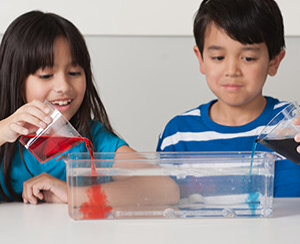 Two children pouring liquids into a glass pan.