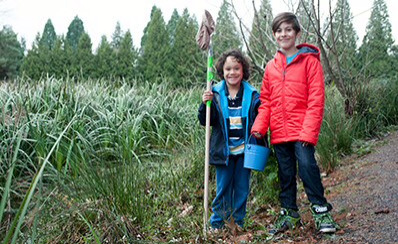 Two young people outdoors carrying a bucket and a net for a science experiment.