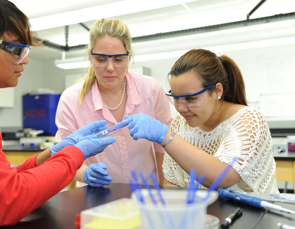 A teacher and two students work together during a science lab experiment. They are wearing gloves and safety glasses.