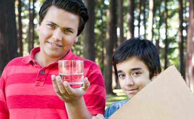 A man lifts up a glass of water while standing next to a child who is holding a piece of cardboard.