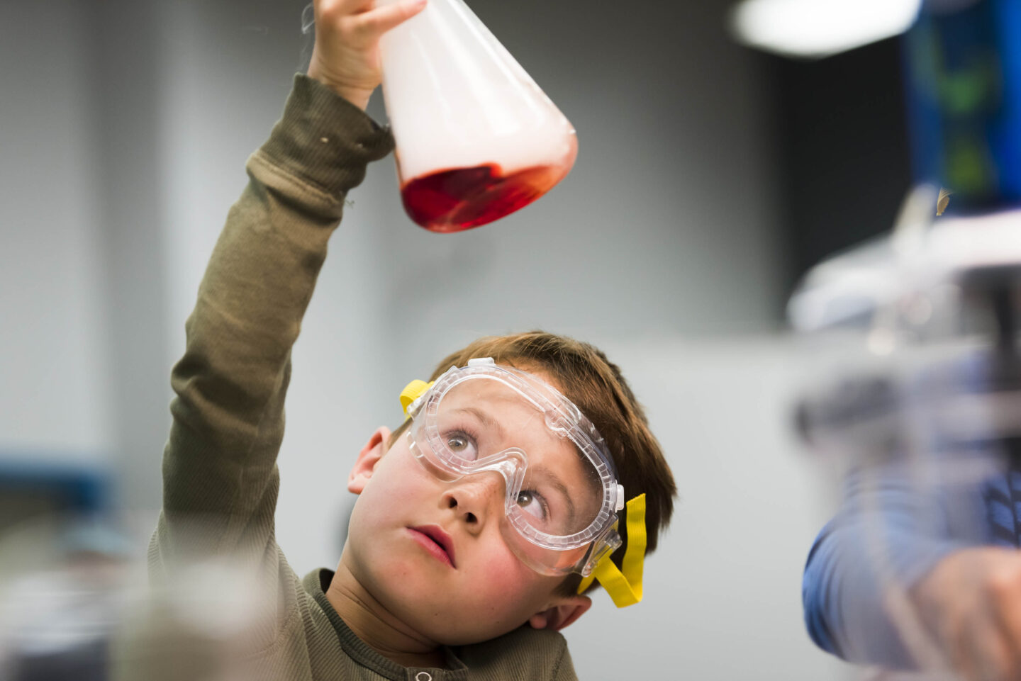 A young person holding up a science beaker with red liquid and wearing goggles