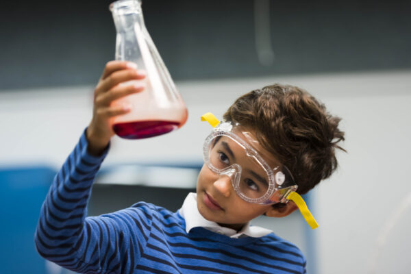 A young person wearing safety googles is holding up a science beaker that contains red liquid