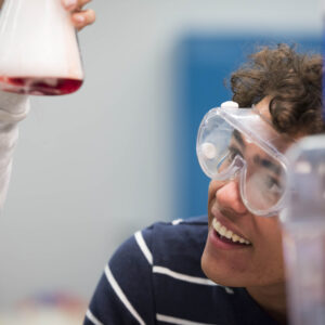 A student wearing safety goggles is looking at a science beaker with red liquid.