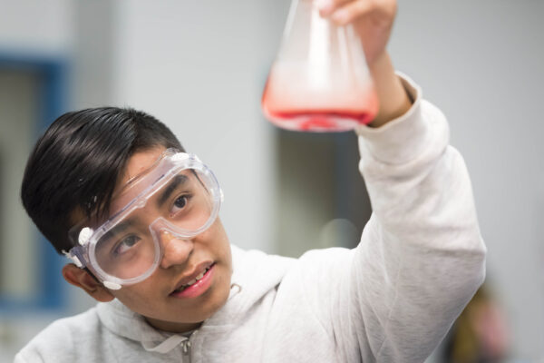 A student is wearing safety goggles and holding up a science beaker with red liquid.