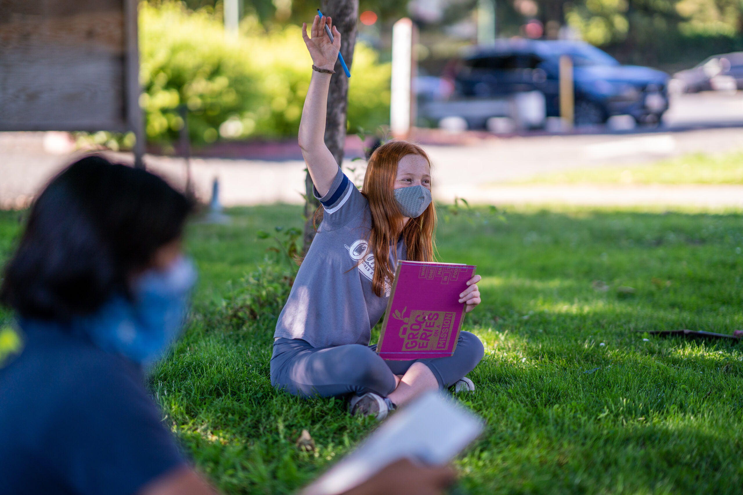 Masked student seated outdoors on a lawn raises hand