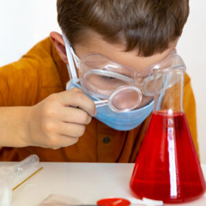 A child wearing safety goggles is looking at red liquid in a science beaker through a magnifying glass