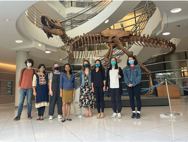 Priya Moorjani in a group photo, standing in front of a skeleton of a dinosaur