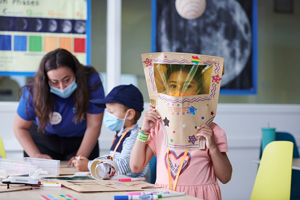 A child wears an astronaut helmet she has constructed during the Moon Explorers camp while another child and camp leader create their own