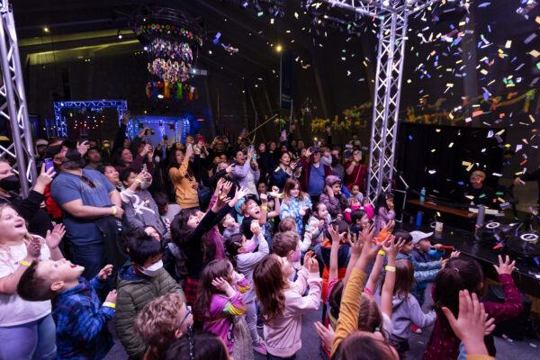 A lively crowd of children and adults watches confetti fall, with many reaching up in excitement.