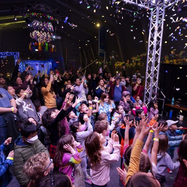 A lively crowd of children and adults watches confetti fall, with many reaching up in excitement.