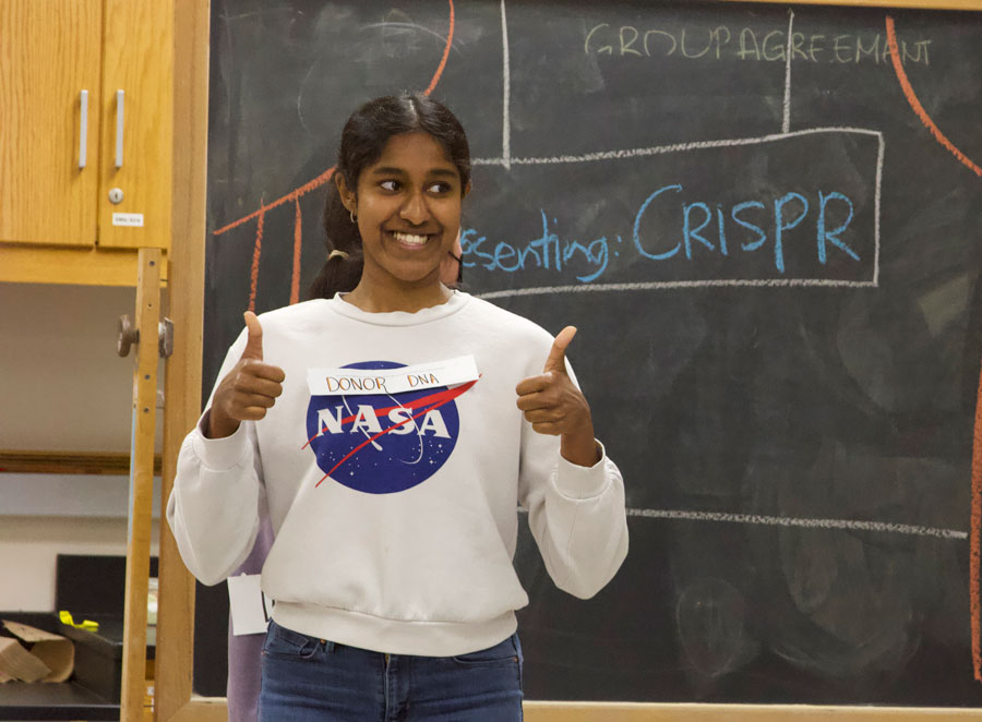 A student stands at a chalkboard and smiles and gives a thumbs up.