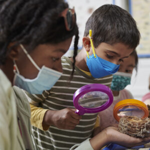 A summer camp educator and a young boy examine a bird nest using magnifying glasses.