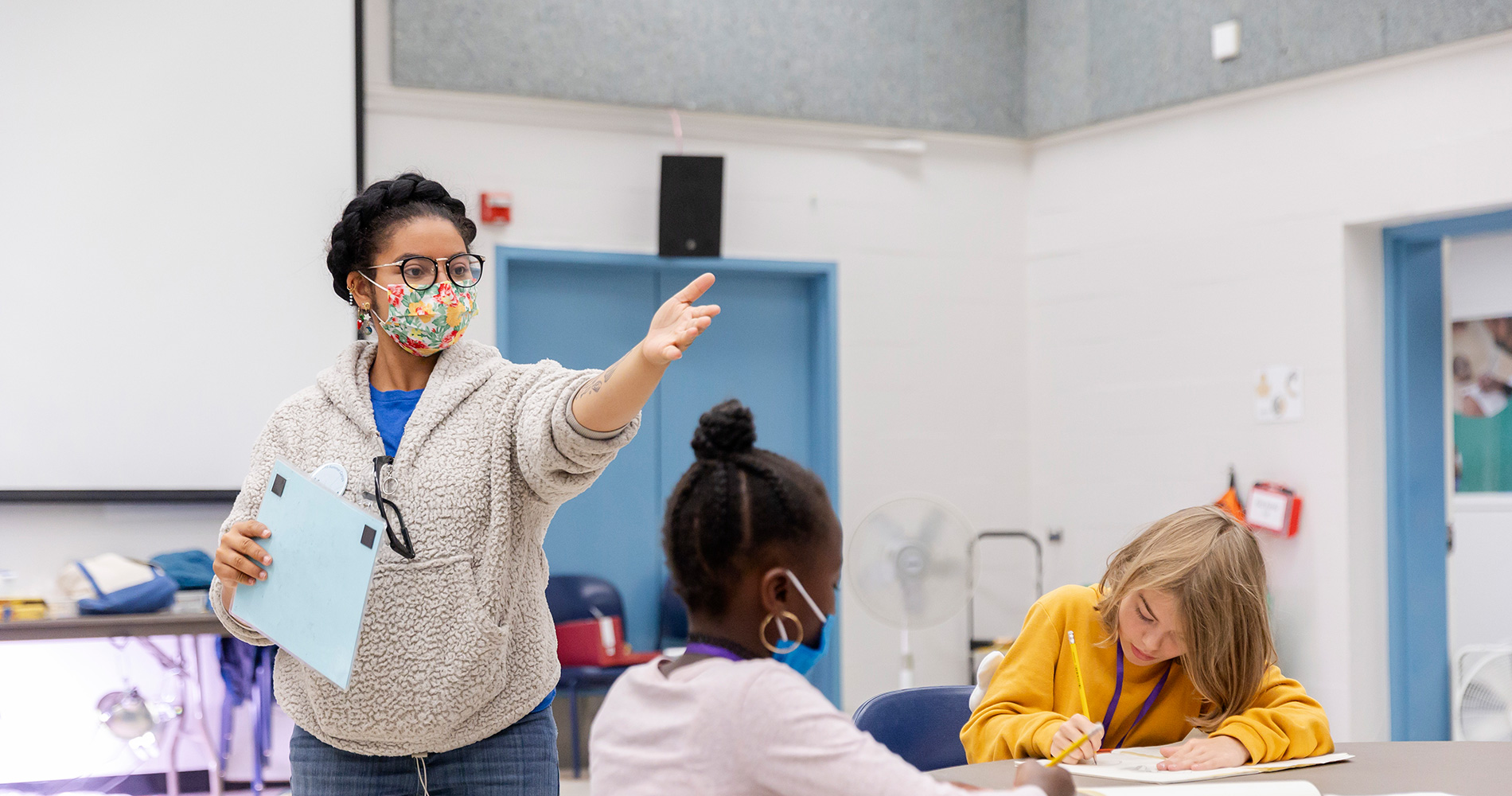 A summer camp educator is facilitating a classroom activity. Two students are seated at a table.