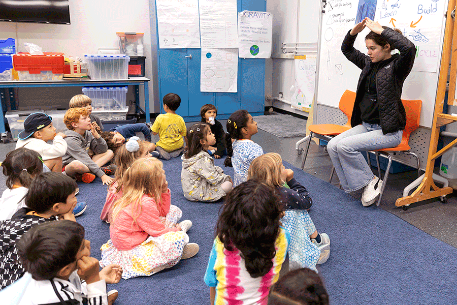 A summer camp educator is leading a classroom activity, her hands raised above her head, and young children are gathered around and seated on the classroom floor.