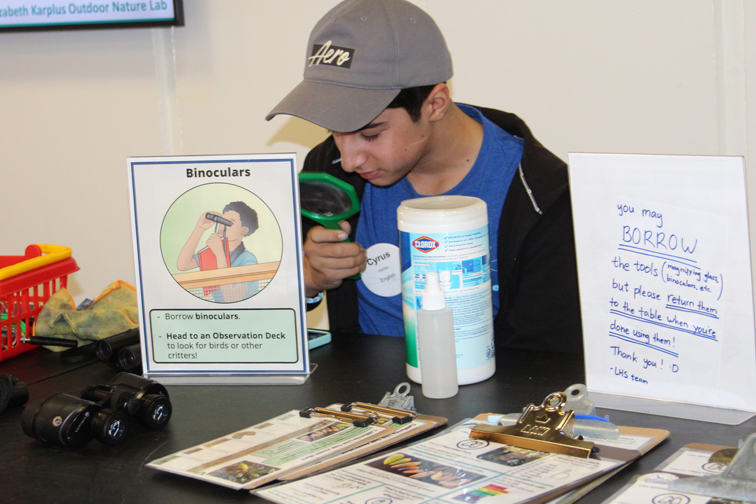 A teen volunteer is looking through a hand lens at a booth with a science activity