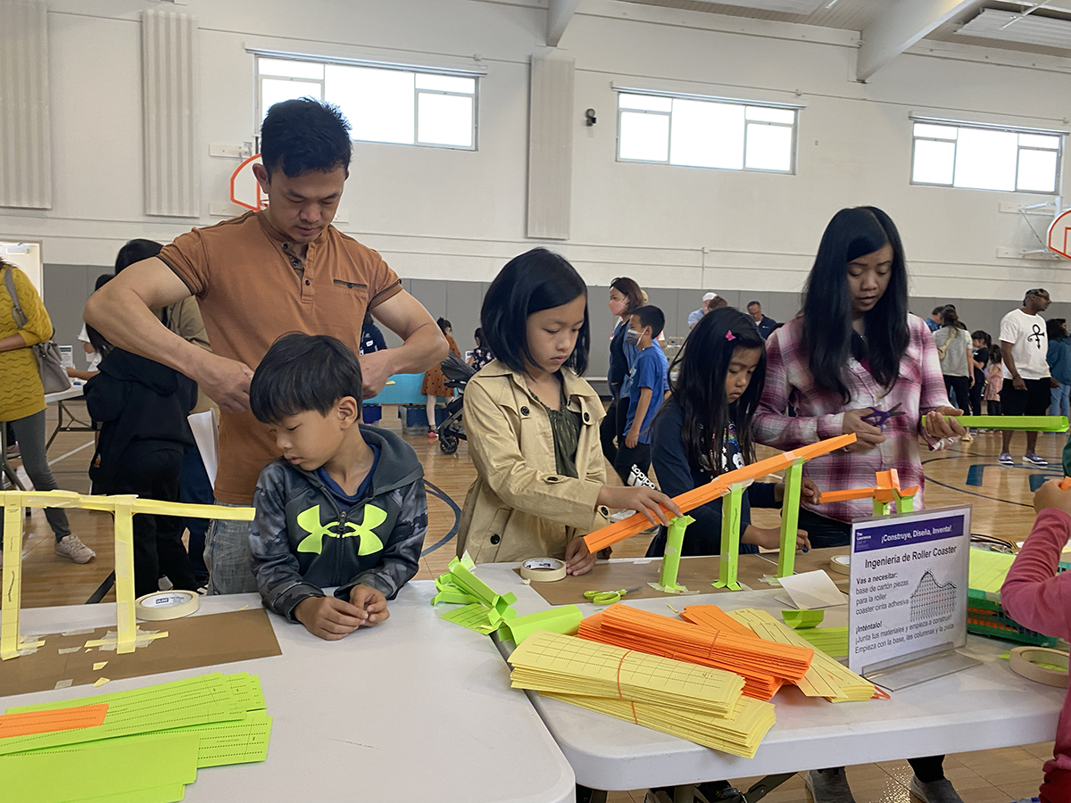 A family building a miniature roller coaster using recycled materials during a Build, Engineer, Invent science festival.