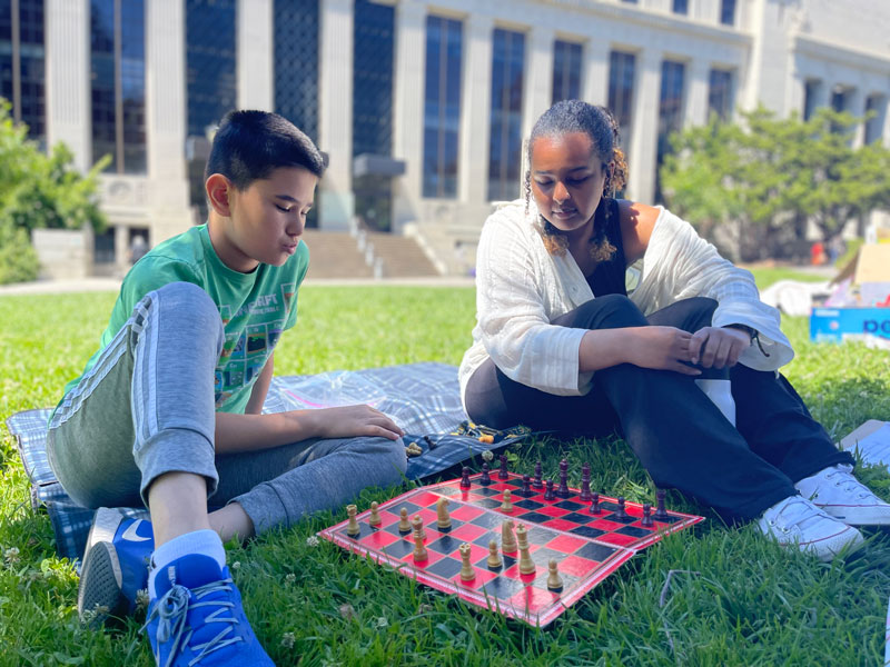 A mentor is mentoring a teen while they play a chess game together on campus