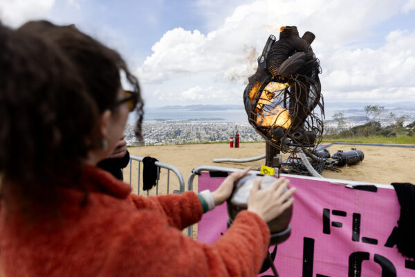 A person in an orange jacket activates a fiery metal heart sculpture on a hilltop.