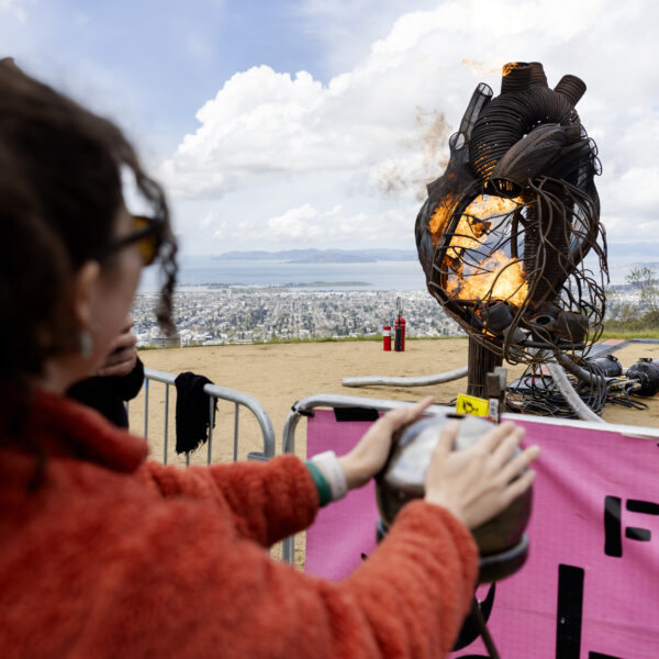 A person in an orange jacket activates a fiery metal heart sculpture on a hilltop.