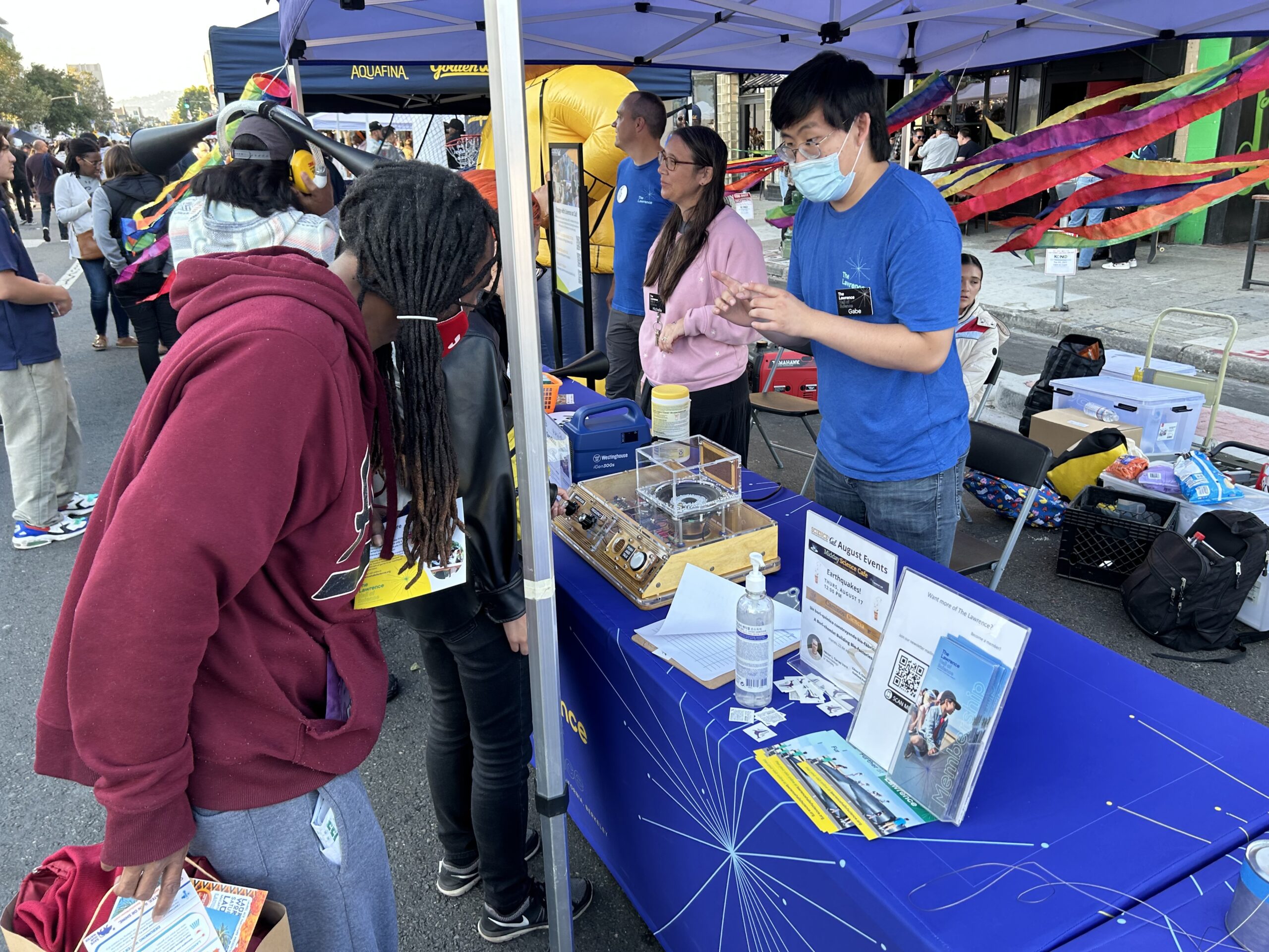 A person is examining an insect display under a magnifying glass at a blue-covered table, while another person in a blue mask explains.