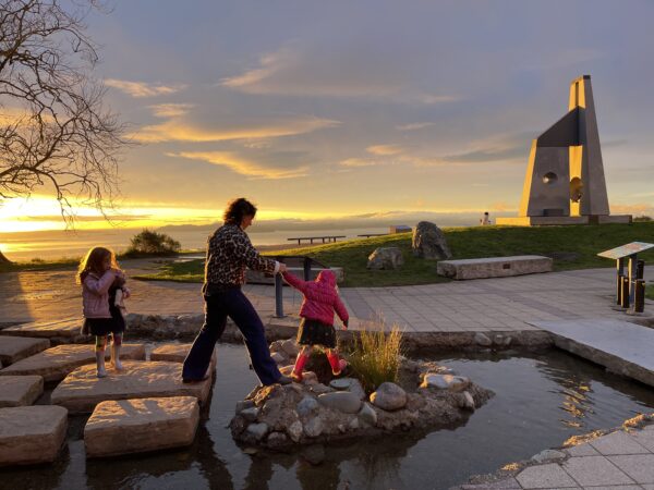 A parent helps a child across stepping stones at a park, with another child watching as the sun sets in the background.