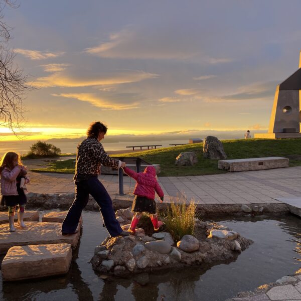 A parent helps a child across stepping stones at a park, with another child watching as the sun sets in the background.