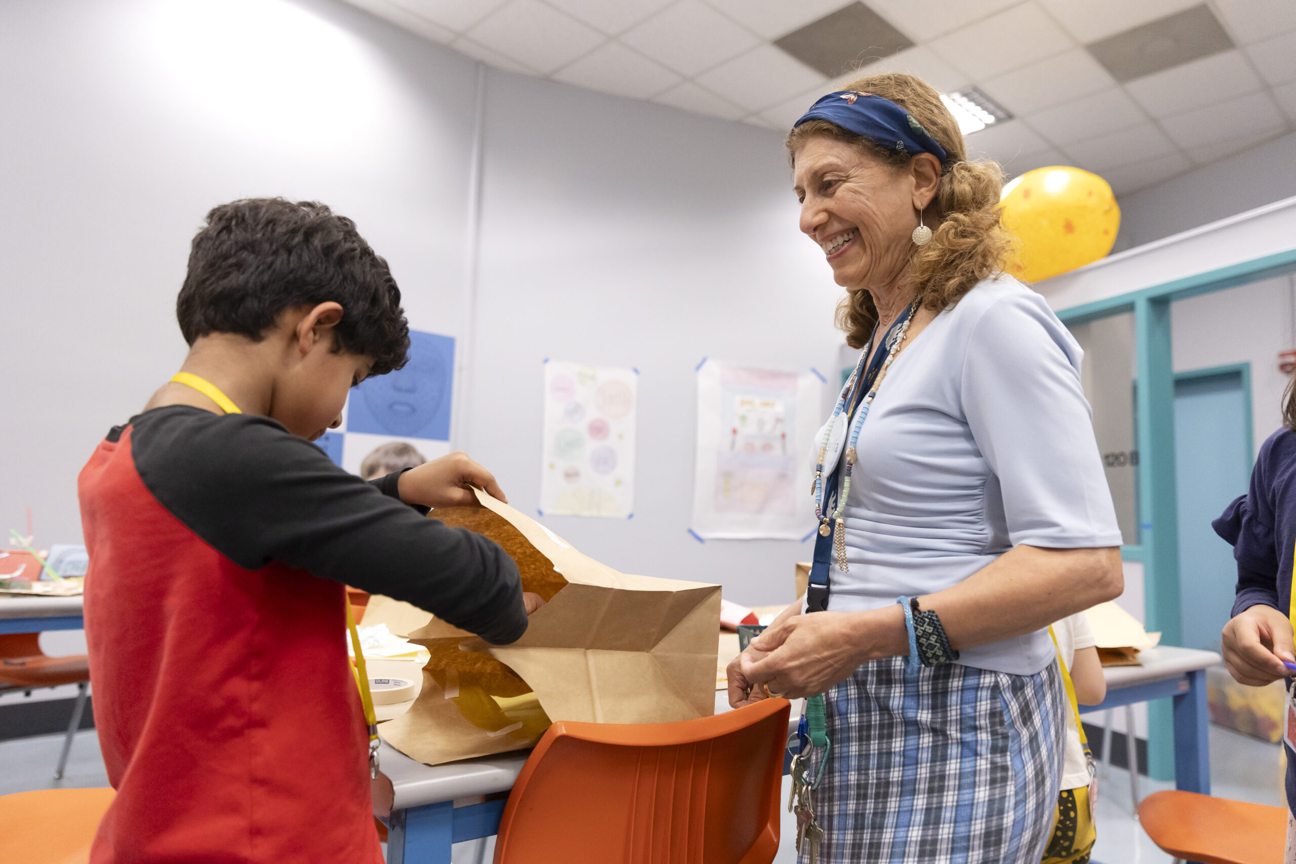 A woman with curly hair and a blue headband smiles as she watches a boy in a red shirt examine the contents of a brown paper bag in a brightly lit classroom.