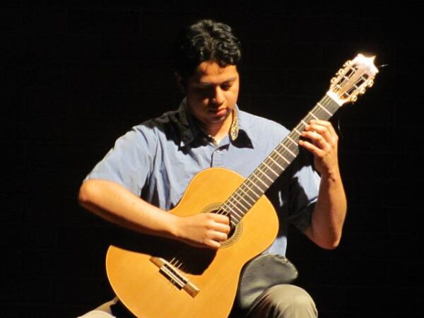 Close-up of a musician concentrating while strumming an acoustic guitar, wearing a blue shirt, with a dark, shadowy backdrop.