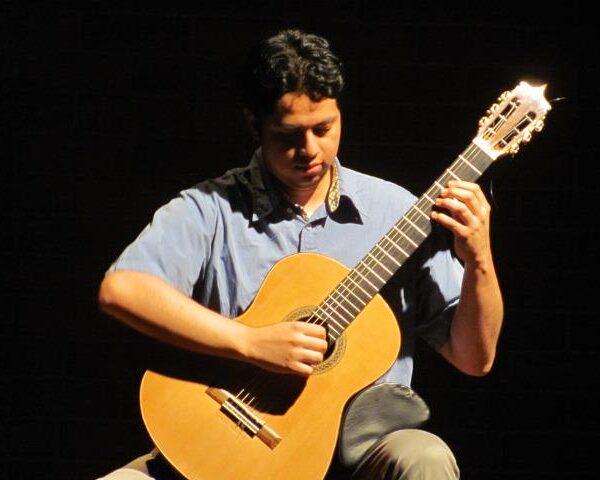 Close-up of a musician concentrating while strumming an acoustic guitar, wearing a blue shirt, with a dark, shadowy backdrop.