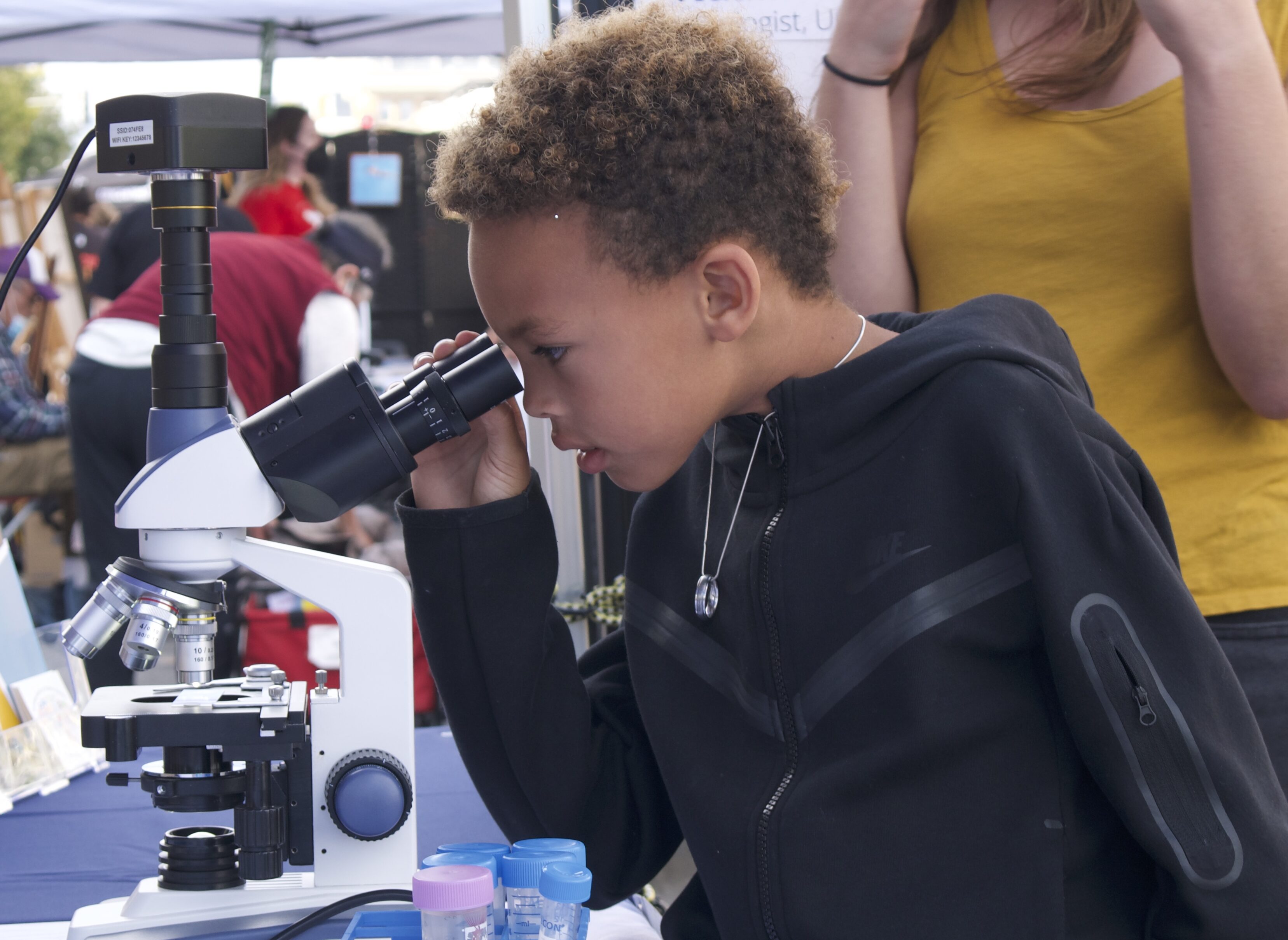 A child is looking through a microscope at an outdoor science event.