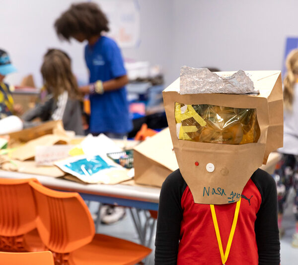 A young child wears an astronaut helmet created during camp while children and their camp leader construct helmets in the classroom.