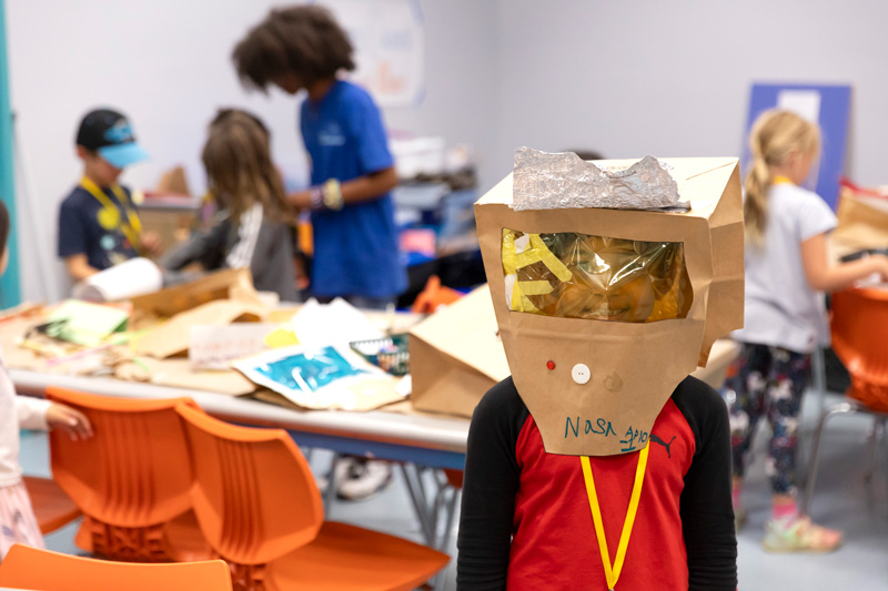 A young child wears an astronaut helmet created during camp while children and their camp leader construct helmets in the classroom.