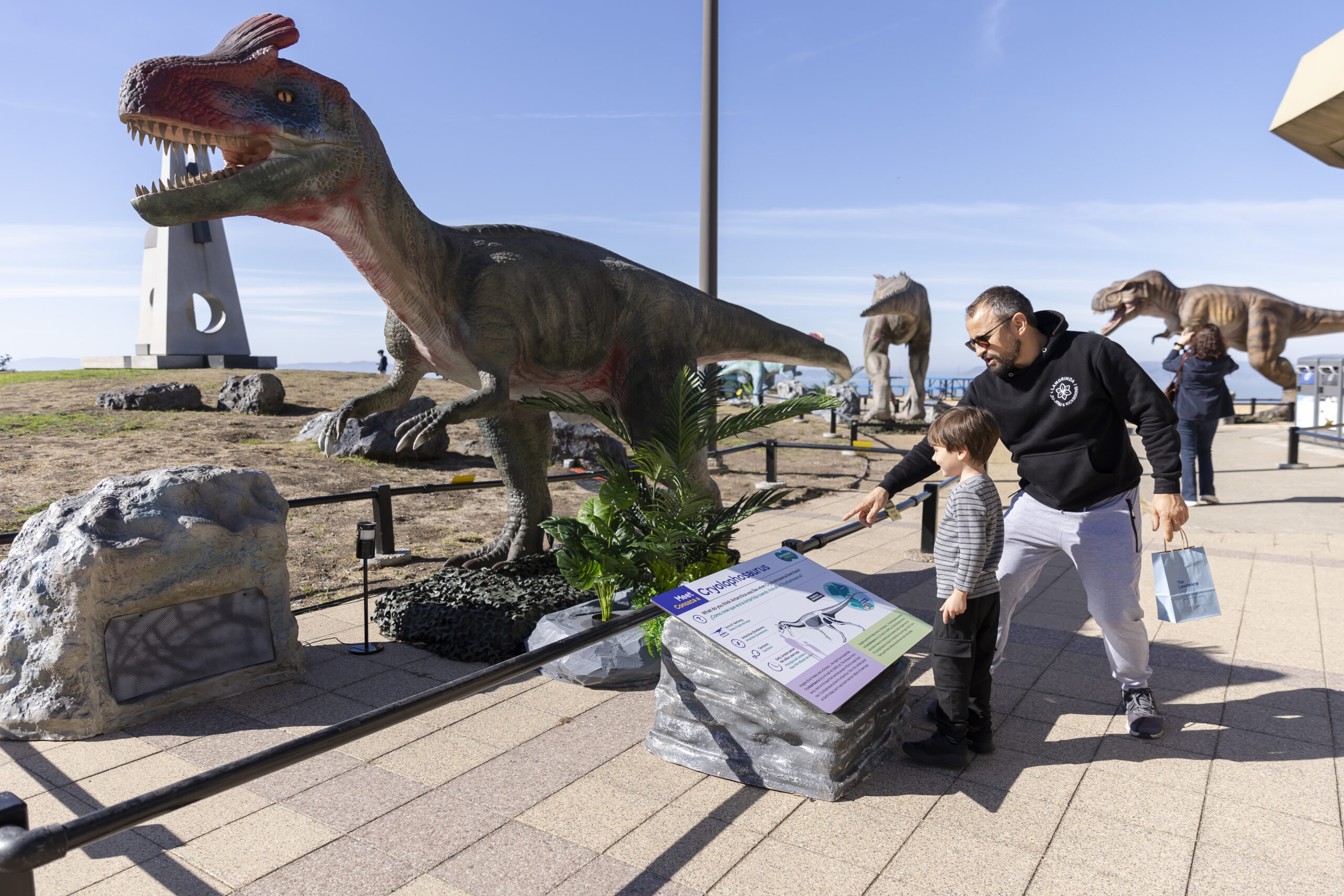 Adult and child exploring a dinosaur exhibit outdoors, with a large model of a Tyrannosaurus Rex and other dinosaur figures in the background.