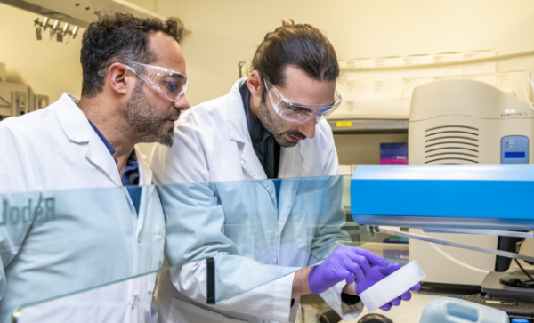 Two scientists in lab coats and safety glasses are working together in a laboratory, examining a sample next to a piece of equipment.