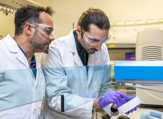 Two scientists in lab coats and safety glasses are working together in a laboratory, examining a sample next to a piece of equipment.