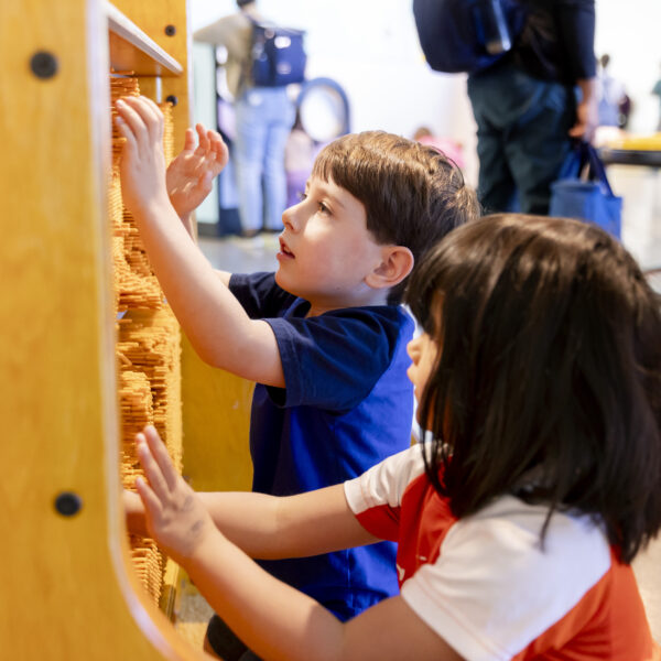 Two children interact with a large tactile wall filled with movable orange pegs in a brightly lit play area.