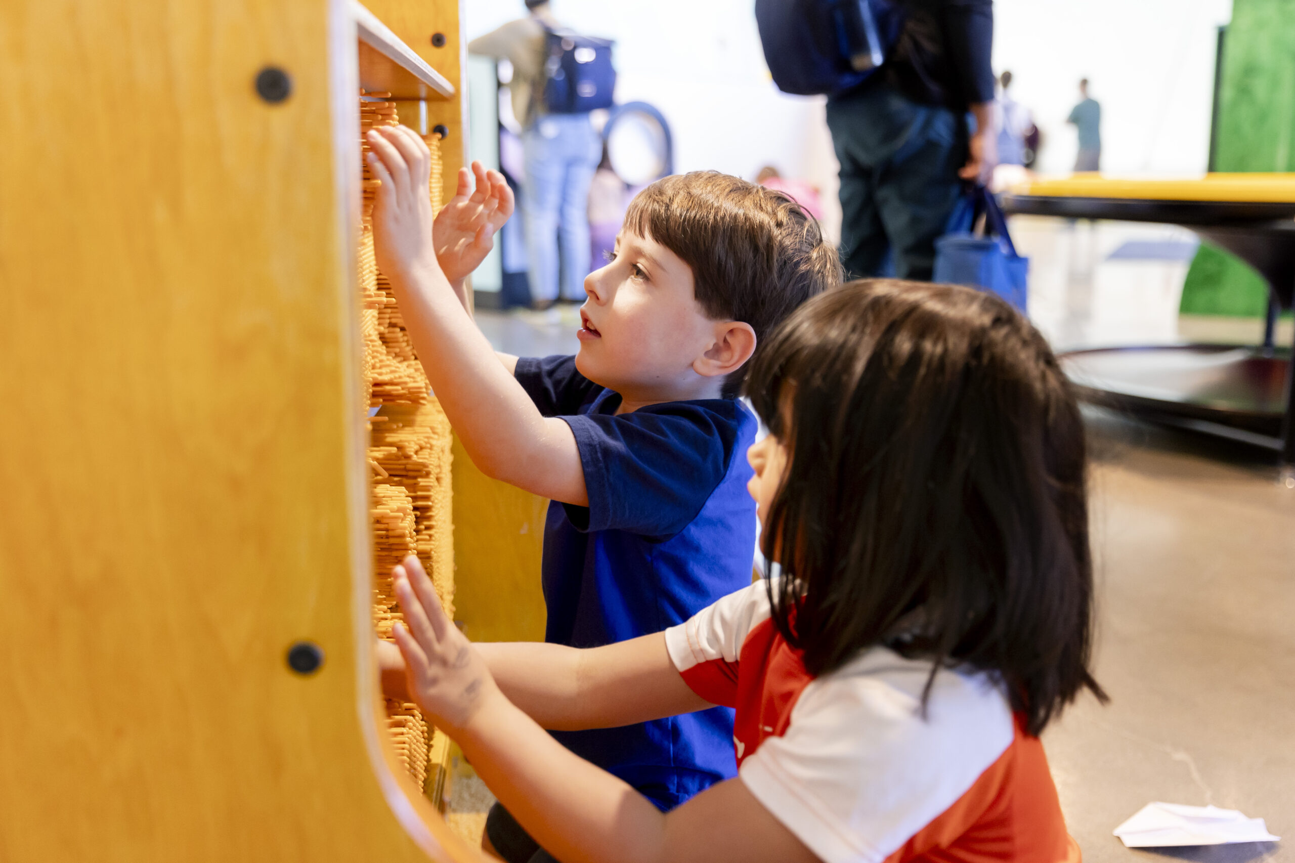 Two children interact with a large tactile wall filled with movable orange pegs in a brightly lit play area.