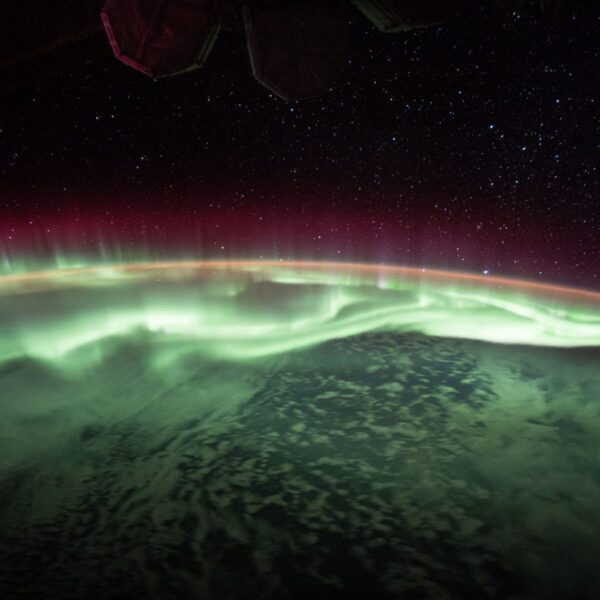 A vibrant green and red aurora spans the curvature of the Earth, as seen from the International Space Station