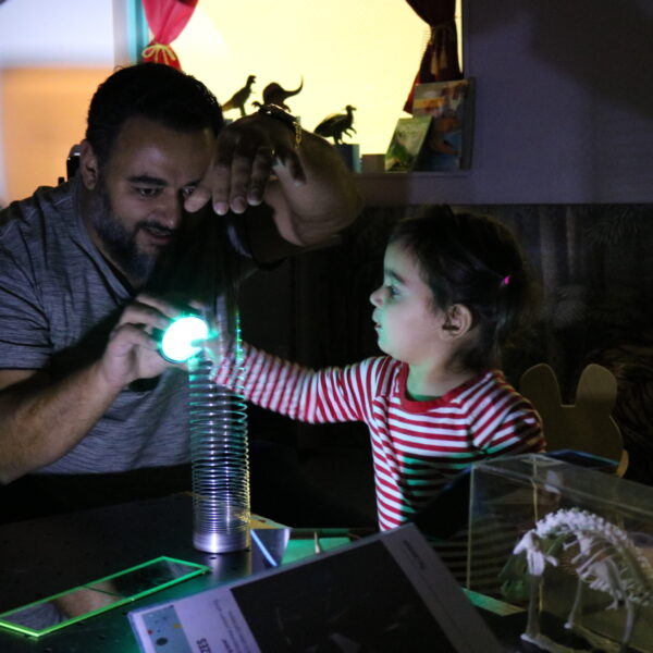 An adult and a child are engaging with a hands-on science exhibit in a dimly lit room, exploring a glowing object together.