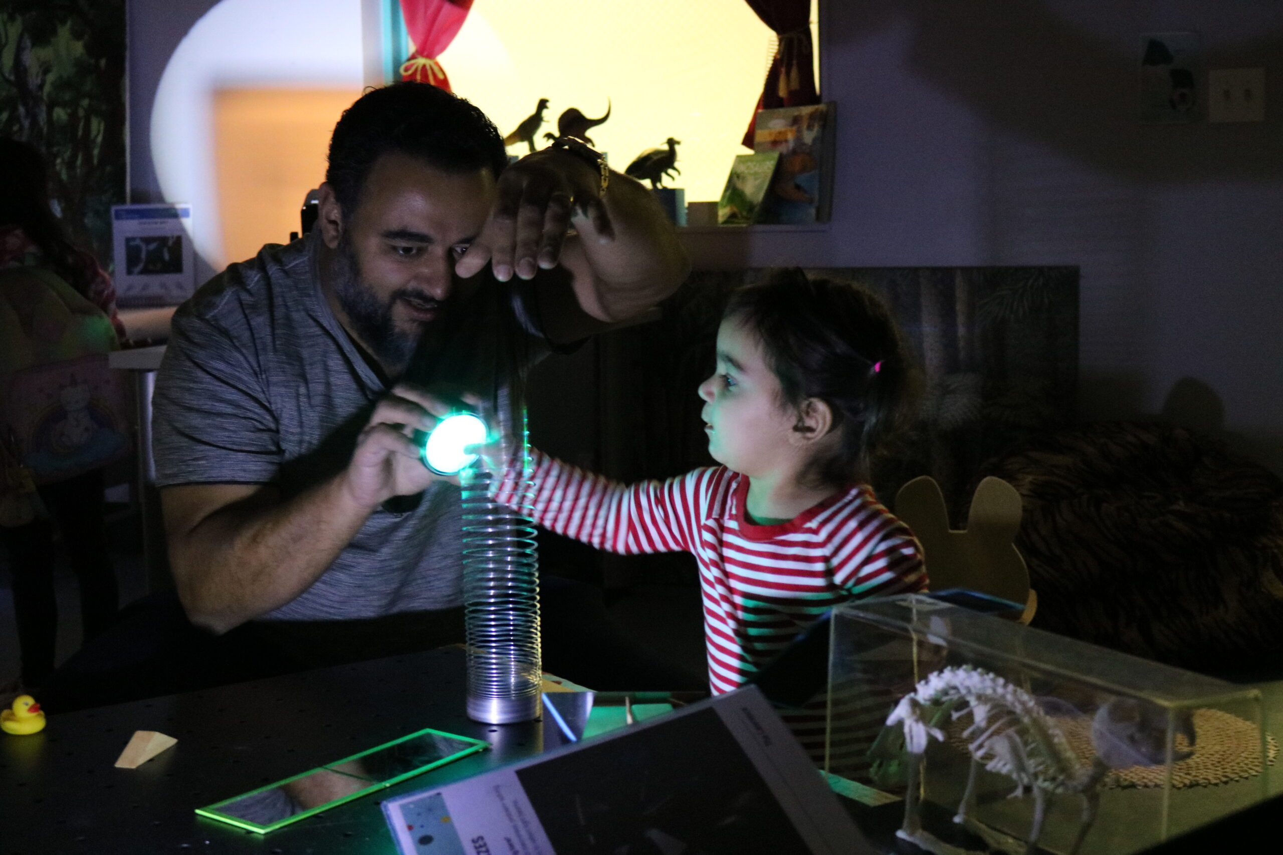 An adult and a child are engaging with a hands-on science exhibit in a dimly lit room, exploring a glowing object together.