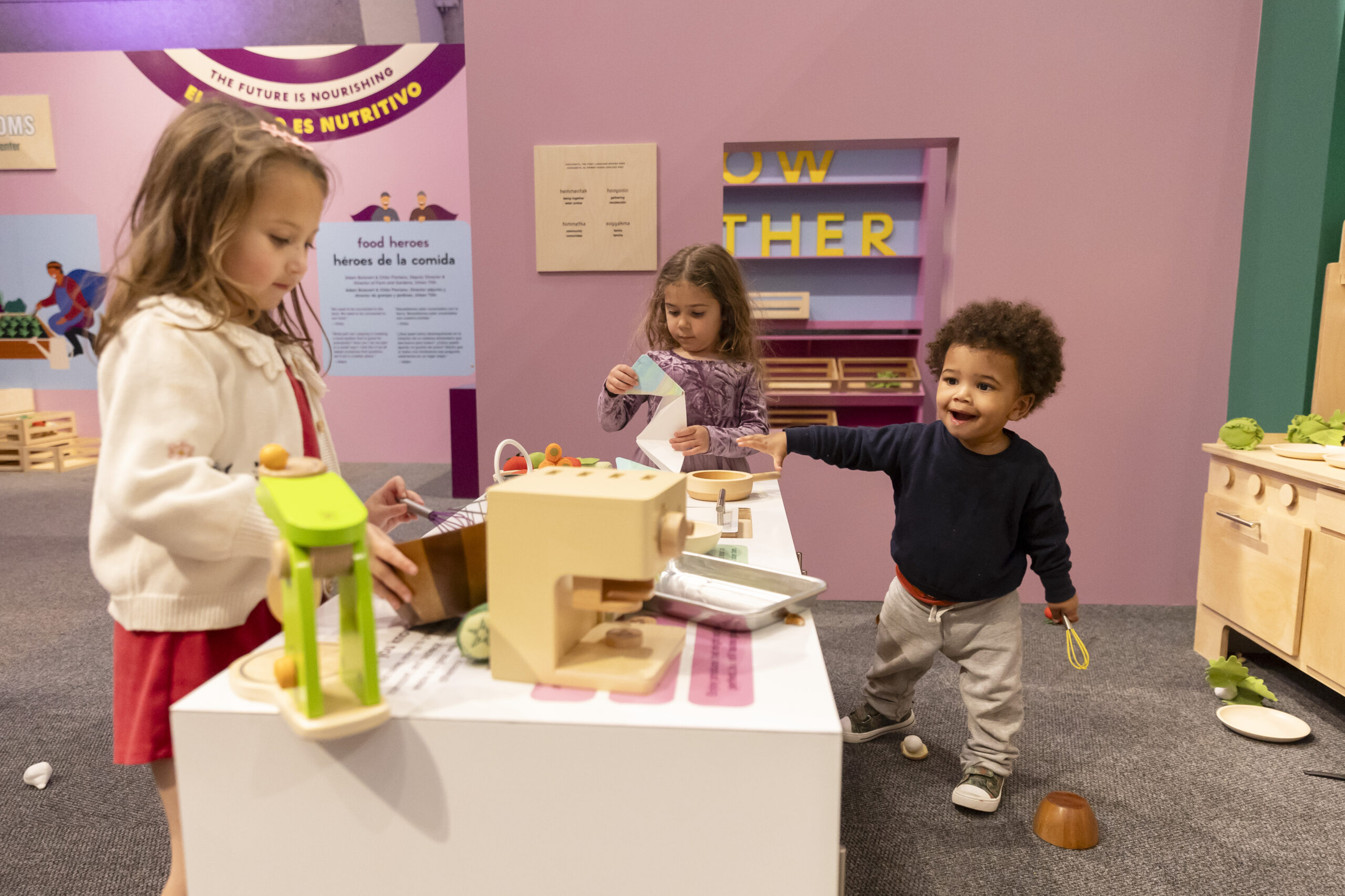 hree children engaging in play at an interactive exhibit featuring a pretend kitchen setup.