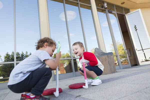 Two children playing launch rockets in front of a glass building under a clear sky.