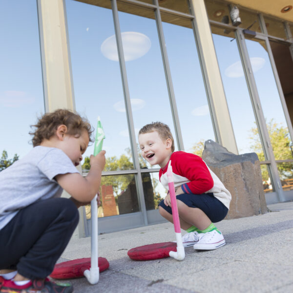 Two children playing launch rockets in front of a glass building under a clear sky.