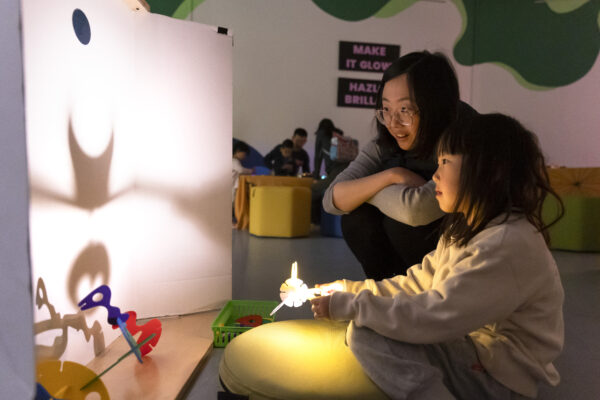 Two individuals engage in a shadow play activity using cut-out figures and a light source to project images on a screen, in a room with others in the background.
