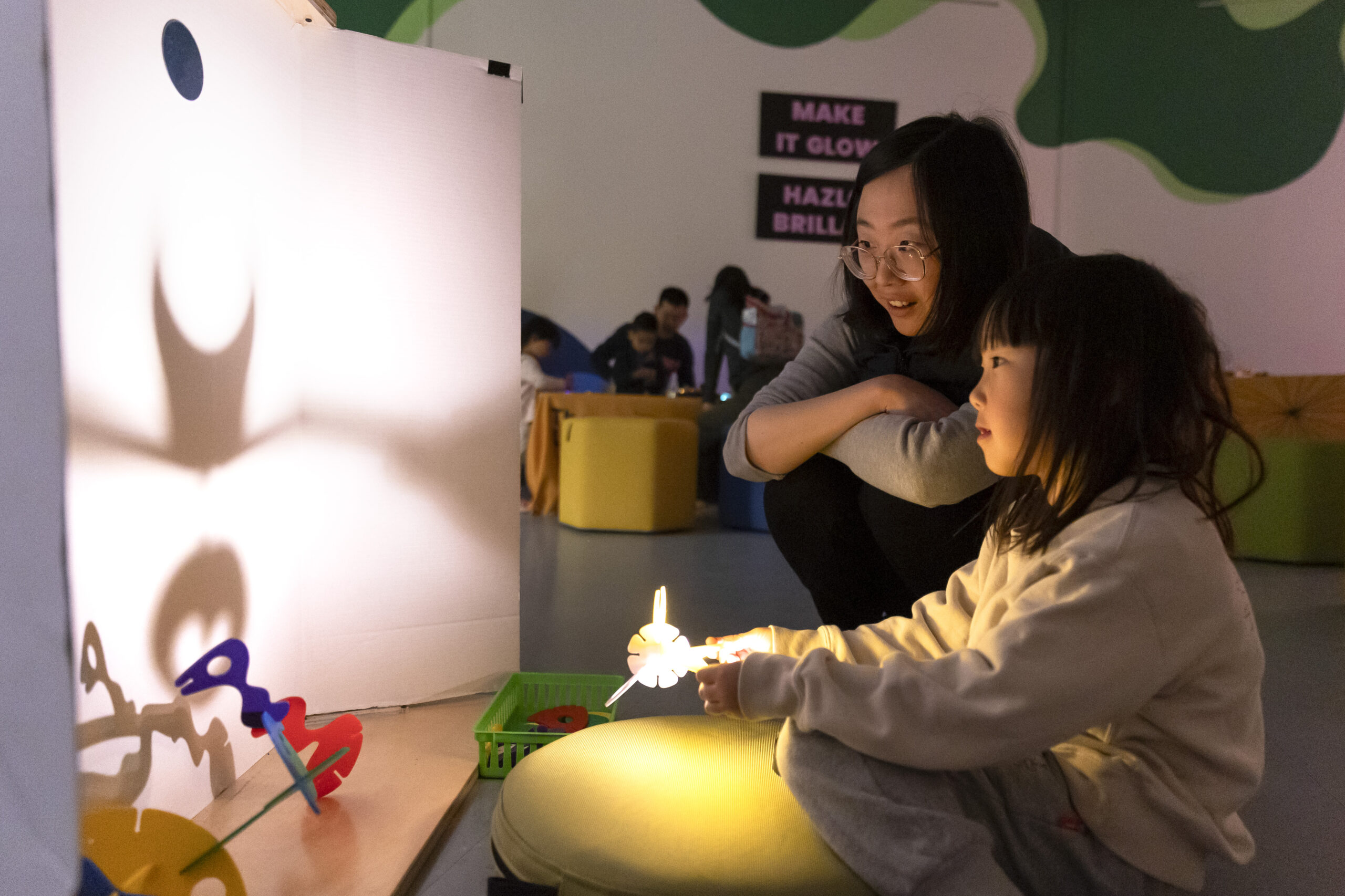Two individuals engage in a shadow play activity using cut-out figures and a light source to project images on a screen, in a room with others in the background.