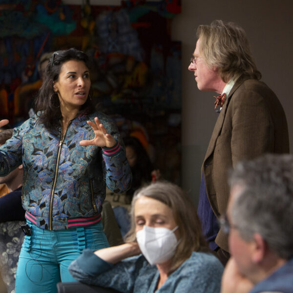 Two individuals engaged in a conversation in a room with attendees are observing their discussion.