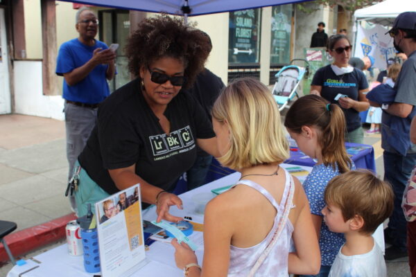 A staff member from The Lawrence discusses with two young visitors at a community event, surrounded by informational pamphlets and books on the table.
