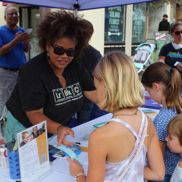 A staff member from The Lawrence discusses with two young visitors at a community event, surrounded by informational pamphlets and books on the table.