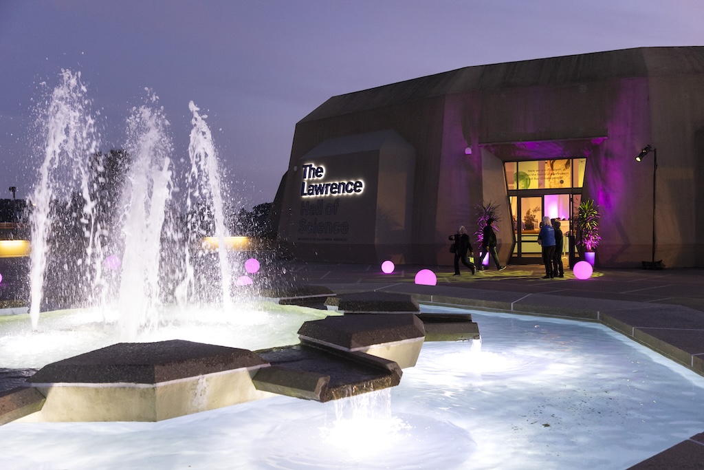 Fountain and purple-lit entrance of The Lawrence Hall of Science at night.
