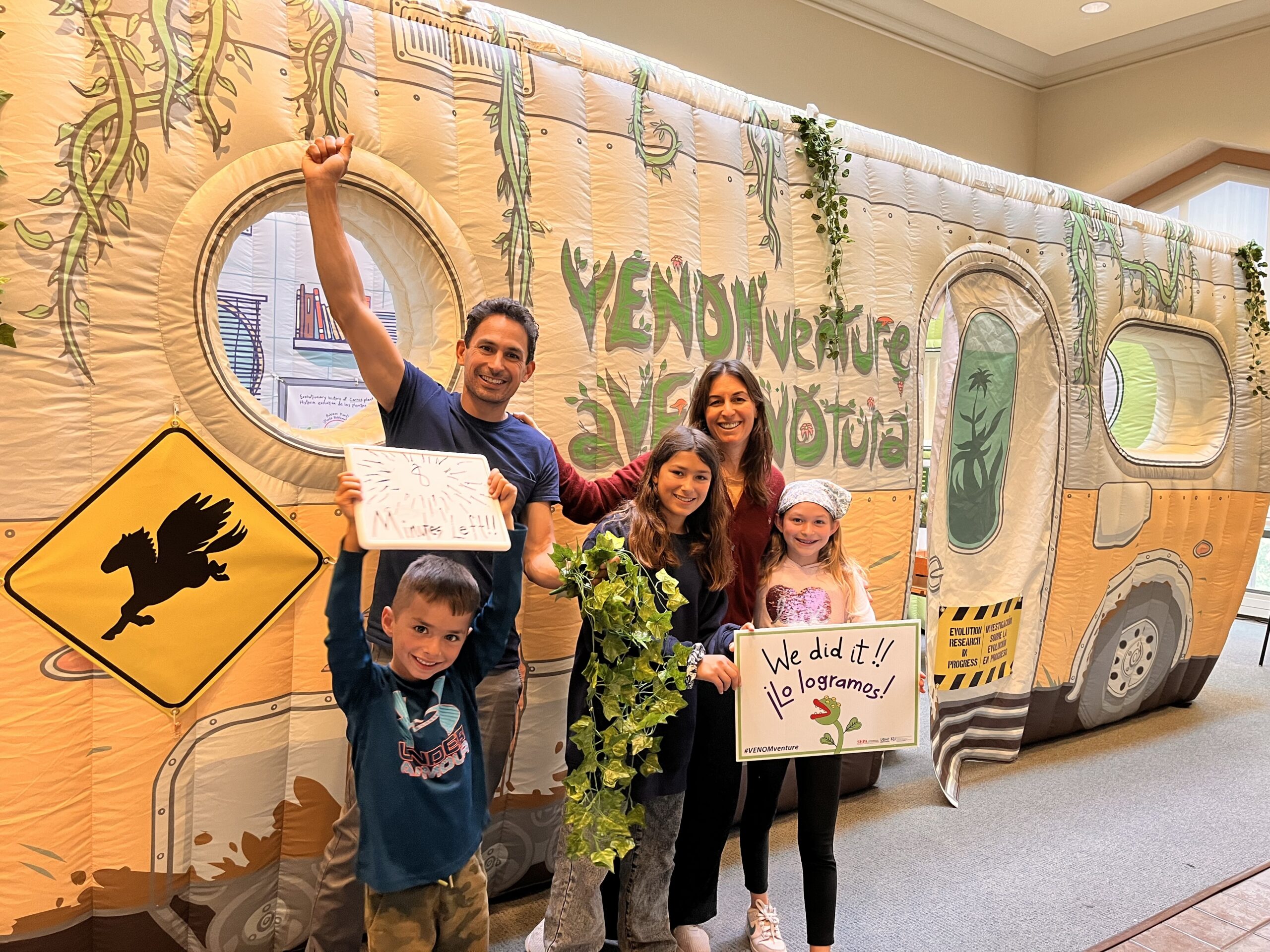 Group of five people, with three adults and two children, posing joyfully in front of a decorative backdrop with a inflatable camper van design and the phrase "Venomventure." They are holding celebratory signs, including one that says "We did it!